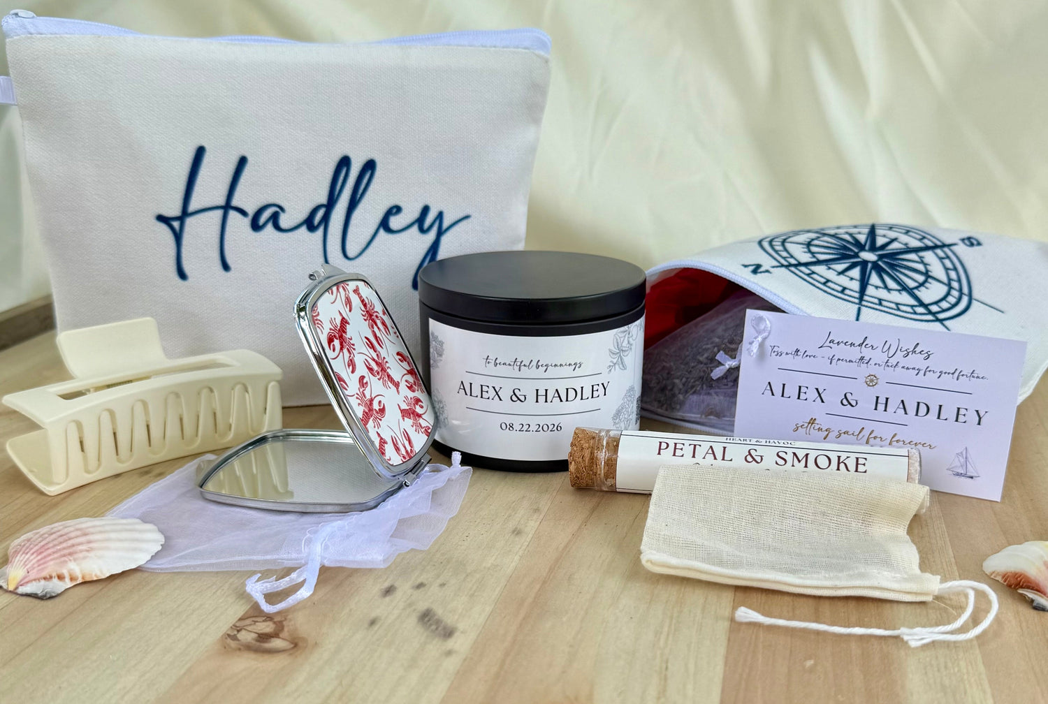 A curated gift set on a light wood surface featuring a personalized cream canvas bag with 'Hadley' embroidered in blue, a scented candle labeled 'ALEX & HadLEY', a lip balm tube labeled 'Petal & Smoke', a small mirror, a compact with a red-and-white floral lid, a drawstring pouch, a hair claw clip, and seashells, all coordinated with matching printed cards and a compass print pouch.