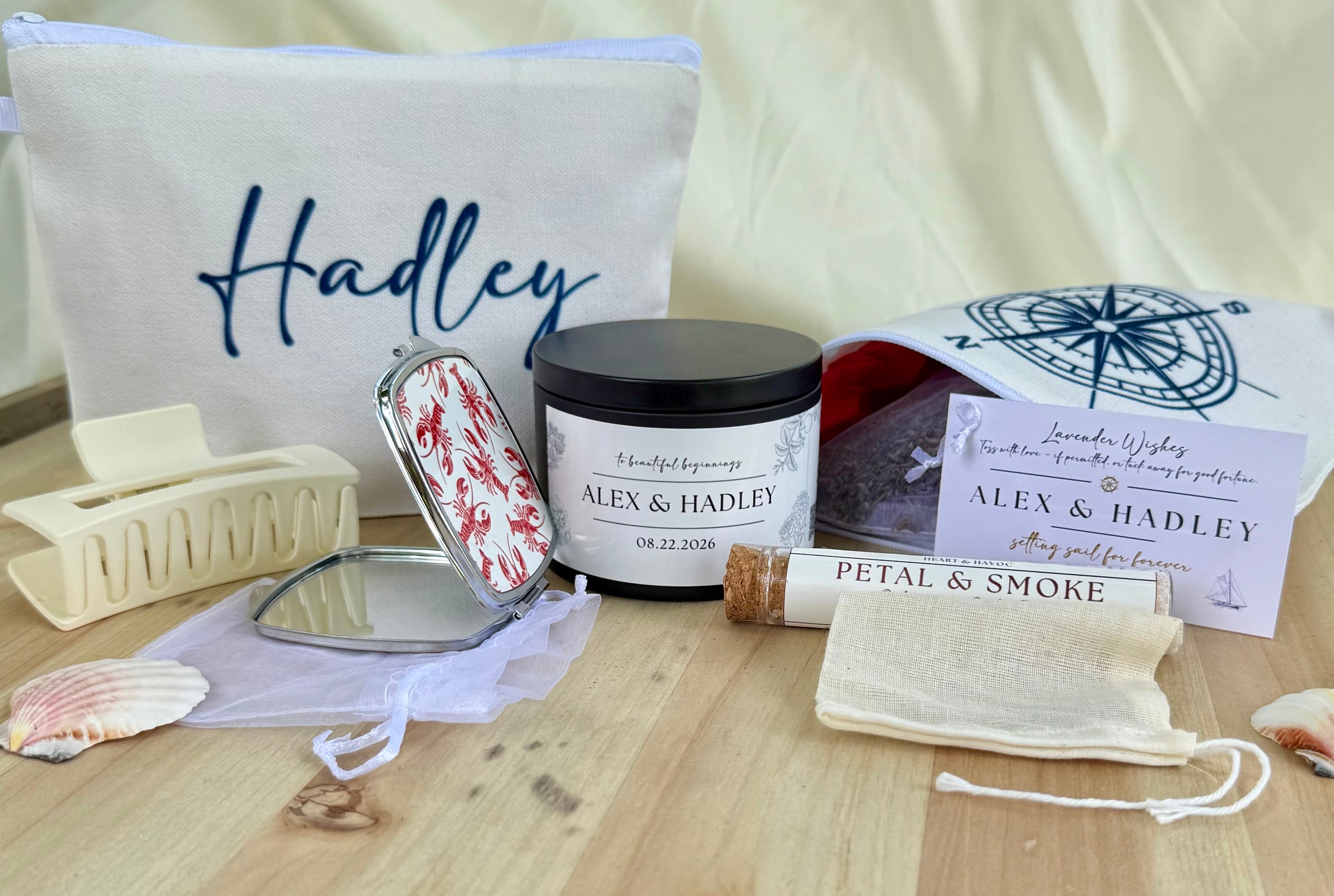 A curated gift set on a light wood surface featuring a personalized cream canvas bag with 'Hadley' embroidered in blue, a scented candle labeled 'ALEX & HadLEY', a lip balm tube labeled 'Petal & Smoke', a small mirror, a compact with a red-and-white floral lid, a drawstring pouch, a hair claw clip, and seashells, all coordinated with matching printed cards and a compass print pouch.