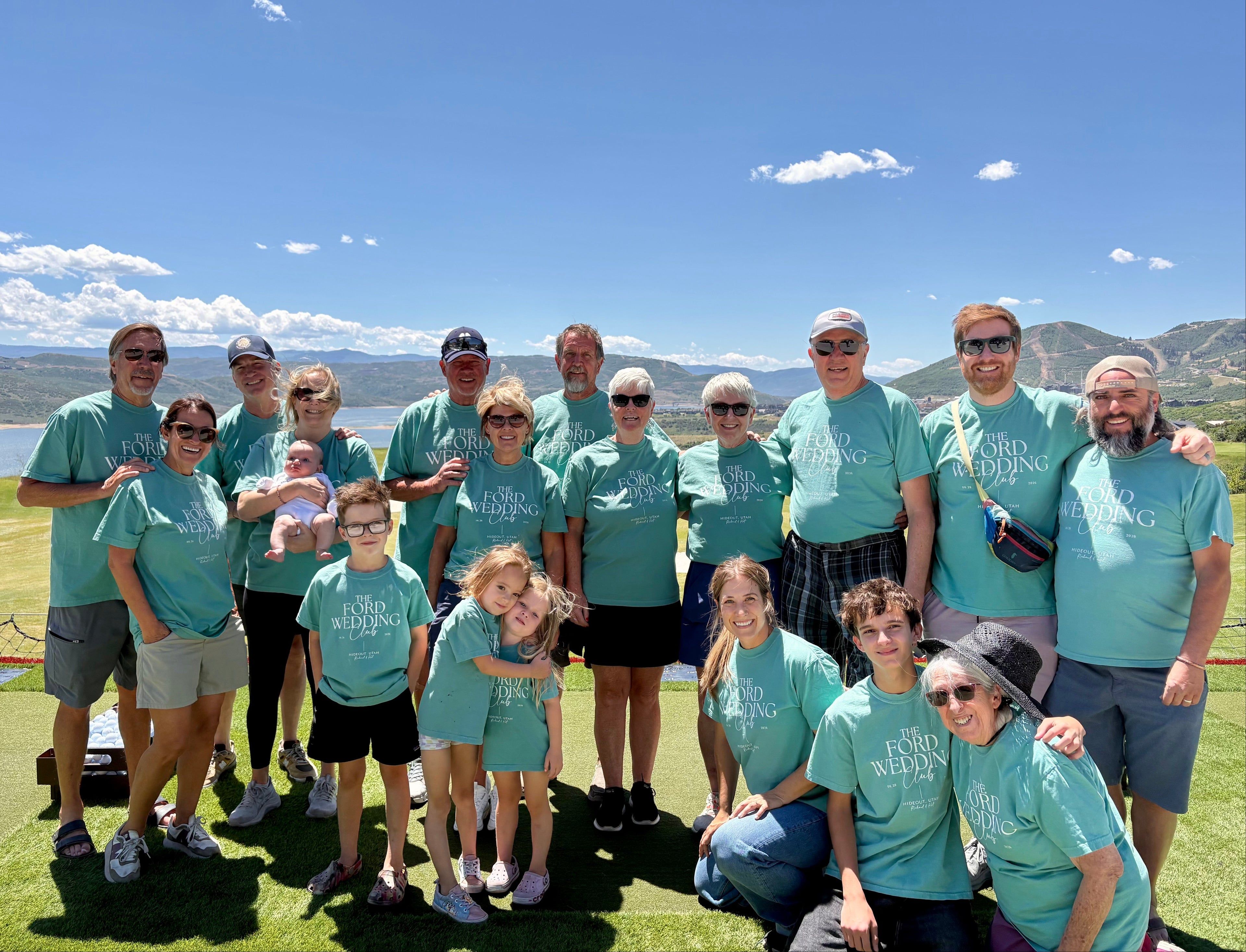 A group of 15 people posing together on a golf course, wearing matching teal t-shirts with 'Spring Weddings' printed in white on the front. The background features a scenic view of a lake, mountains, and a clear blue sky.