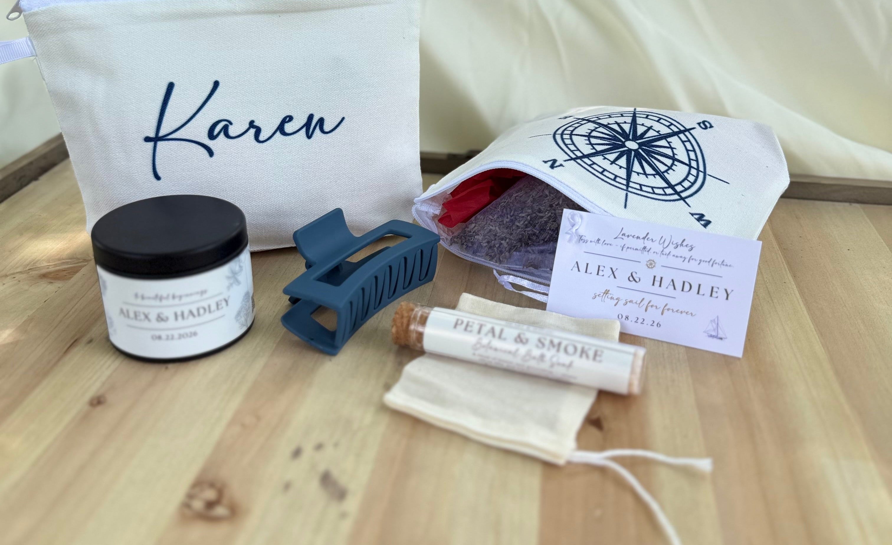 A curated gift set arranged on a light wood tray in front of a cream fabric backdrop. The set includes a white zip pouch with the name Karen in navy script, a small jar labeled SLS & HARLEY, a blue hair clip, a rolled item labeled PEEL & SMORE, a small drawstring pouch, and a printed card with a compass design.