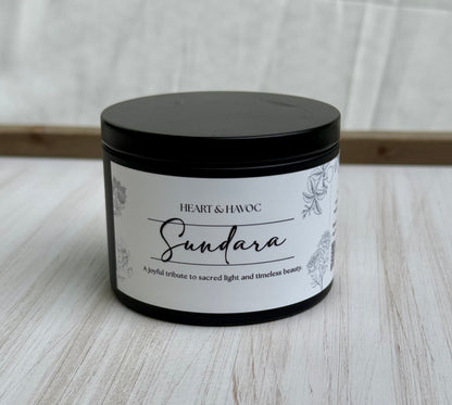 A black metal candle tin with a white label reading 'Heart & Hawc Sundara' on a light wood surface against a white fabric backdrop.
