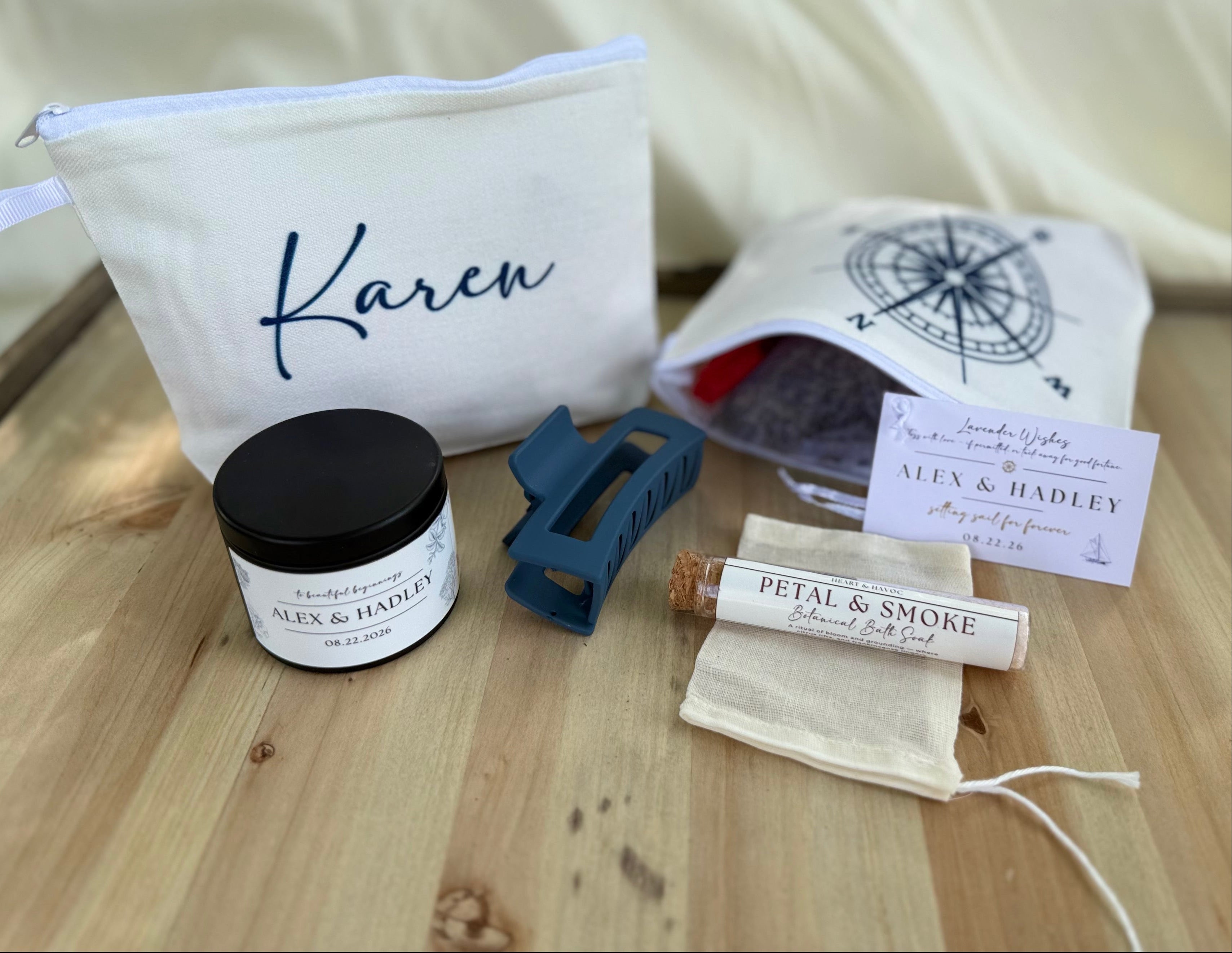 A curated gift set arranged on a light wood surface against a cream fabric backdrop. The set includes a white zip pouch with the name Karen in navy script, a small white jar with a black lid and white label, a blue hair claw clip, a rolled white sachet labeled Petal & Smoke, a cream drawstring pouch, and a printed card.