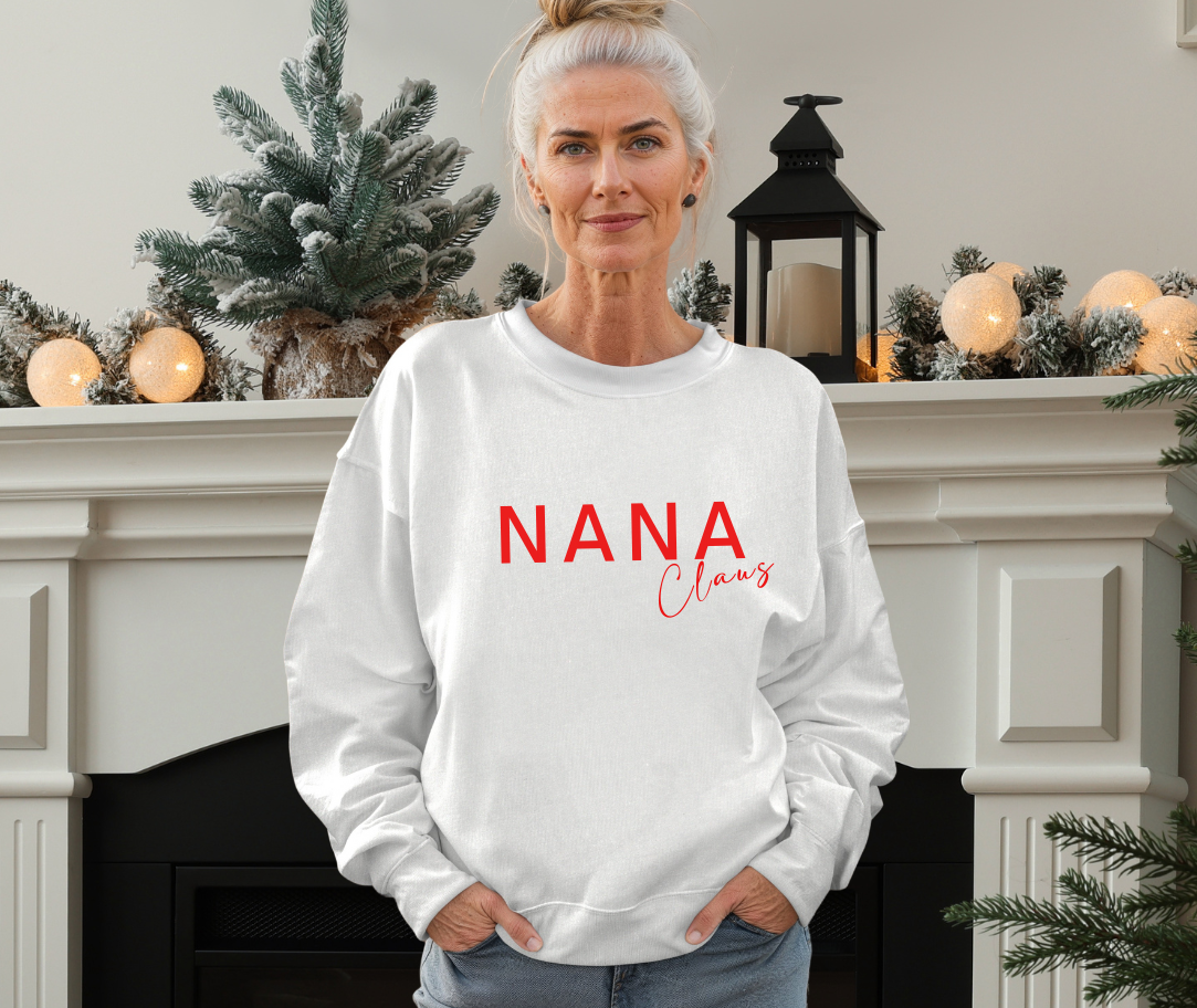 Woman wearing a white sweatshirt with 'NANA Claus' printed in red, standing in front of a decorated fireplace.
