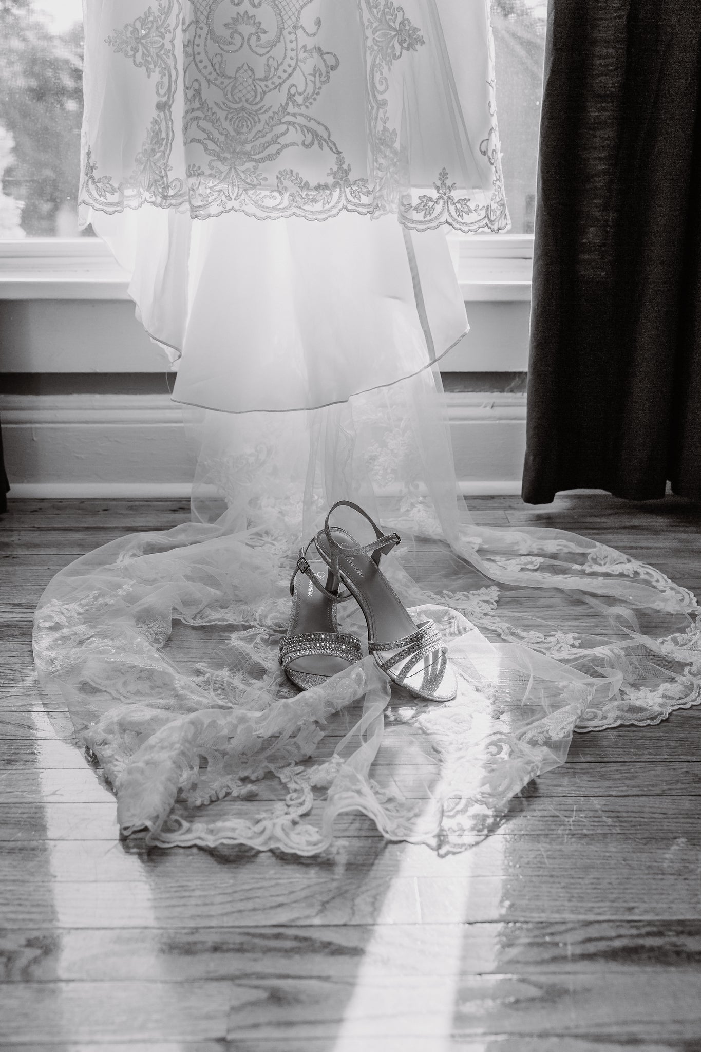 A pair of metallic strappy high-heeled shoes placed on a lace bridal train spread across a wooden floor in front of a window, with a dark curtain on the right.